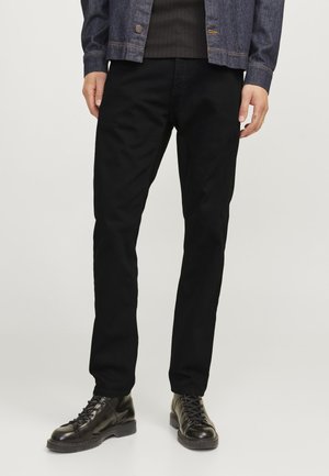 Homme portant un pantalon noir à jambes droites, des bottes en cuir noires à lacets, une chemise côtelée foncée et une veste en denim foncée, debout les mains détendues.