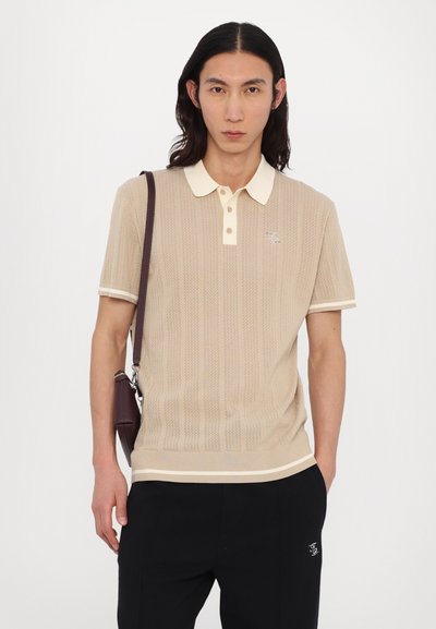 Beige knitted polo shirt with a textured pattern, white collar, and trim. Paired with black trousers and a brown messenger bag.