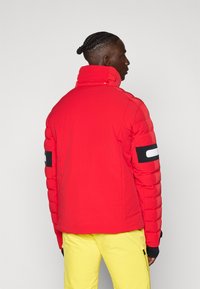 Rote gepolsterte Jacke mit hohem Kragen, ausgestattet mit einem schwarzen Streifen am Ärmel und einer glatten Textur. Kombiniert mit gelben Hosen.