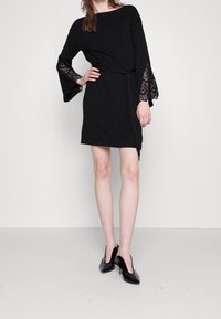 Černé šaty s dlouhými zvonovými rukávy, zdobené krajkovými detaily, s opaskem a rozparkem až po stehna. Nosit s černými lodičkami na vysokém podpatku.