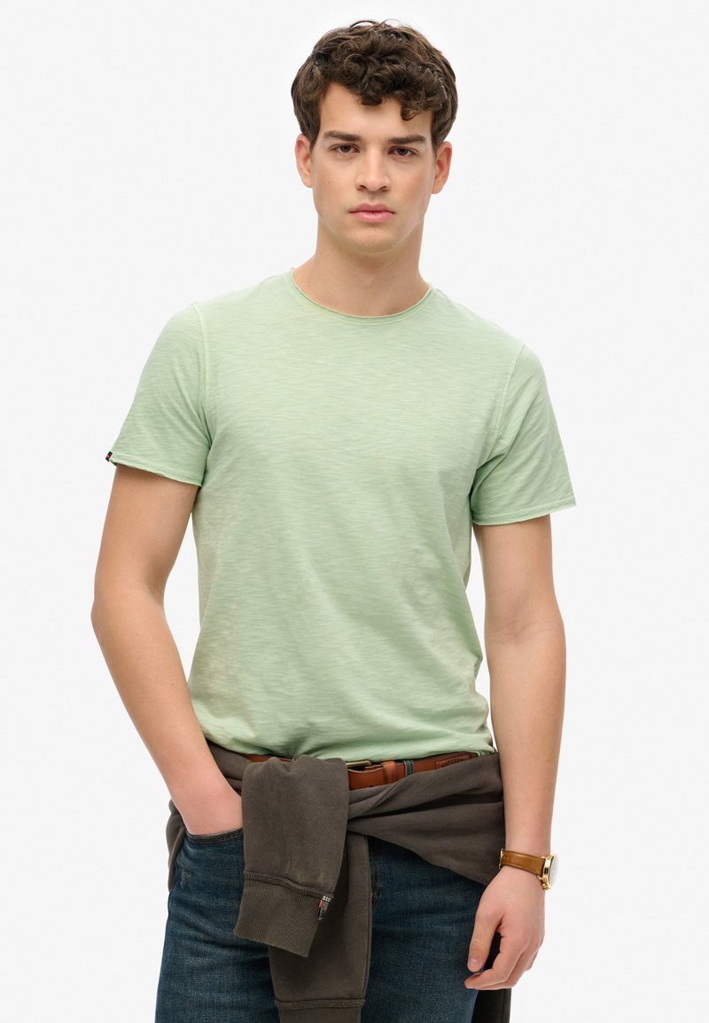 Camiseta de manga corta verde claro con cuello redondo, hecha de material suave. Combinada con jeans oscuros y un suéter marrón atado a la cintura.