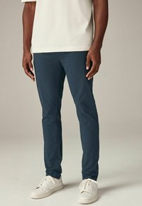 Sötétkék, slim-fit chino nadrág, pamut anyagból készült, sima textúrával, elülső zsebekkel, fehér sportcipőkkel párosítva.
