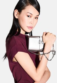 Borse in argento con forma rettangolare, finitura metallizzata e tracolla a catena, tenuta contro un top bordeaux, che mette in mostra linee pulite e un design minimalista.