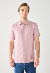 Camisa rosa de botones de manga corta en un tejido texturizado, con cuello y un pequeño logo bordado en el pecho, combinada con jeans azules.