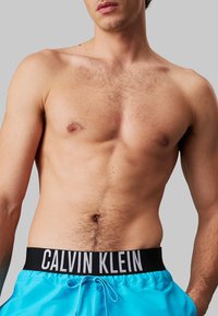 Pantaloni da bagno blu con una vita nera su cui è riportato "CALVIN KLEIN" in bianco. Il tessuto ha una texture liscia e leggera.