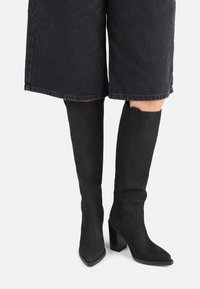 Black knee-high boots made of a smooth material with a block heel. Features a pointed toe and subtle stitching on the sides.