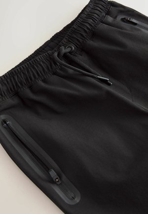 Czarne sportowe szorty z elastycznym paskiem w talii, regulowanym sznurkiem i dwoma bocznymi kieszeniami zapinanymi na zamek. Gładka, lekka tekstura materiału.