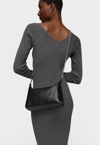 Bolso de cuero negro con textura lisa, con una correa de cadena y un logo sutil, combinado con un vestido gris ajustado de punto acanalado.