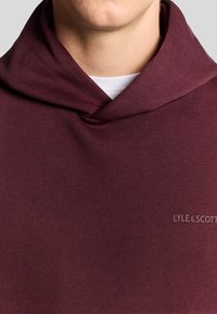 Bordeaux hoodie med en blød tekstur, der har et halsdesign og et diskret logo på den nederste højre side.