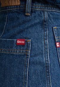 Jeans de mezclilla azul oscuro con costuras naranjas y una etiqueta roja "BIG STAR" en el bolsillo trasero. La tela tiene un acabado texturizado.