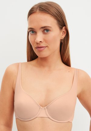 Vrouw met lang lichtbruin haar, een nude-kleurige beha en gouden oorringen tegen een effen achtergrond.