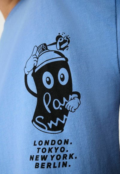 Ljusblå t-shirt med en svart graffitidosa med ögon. Texten nedanför lyder "LONDON. TOKYO. NEWYORK. BERLIN." på framsidan.
