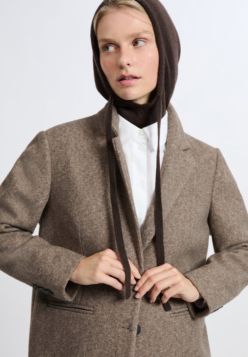 Cappotto di lana marrone con un design su misura, dotato di un colletto, chiusura a bottoni e due tasche. Accompagnato da una sciarpa di lana marrone.