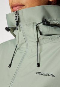 Giacca impermeabile verde chiaro con colletto alto, coulisse nera e chiusura con zip. Presenta un logo sul lato sinistro. Texture liscia.