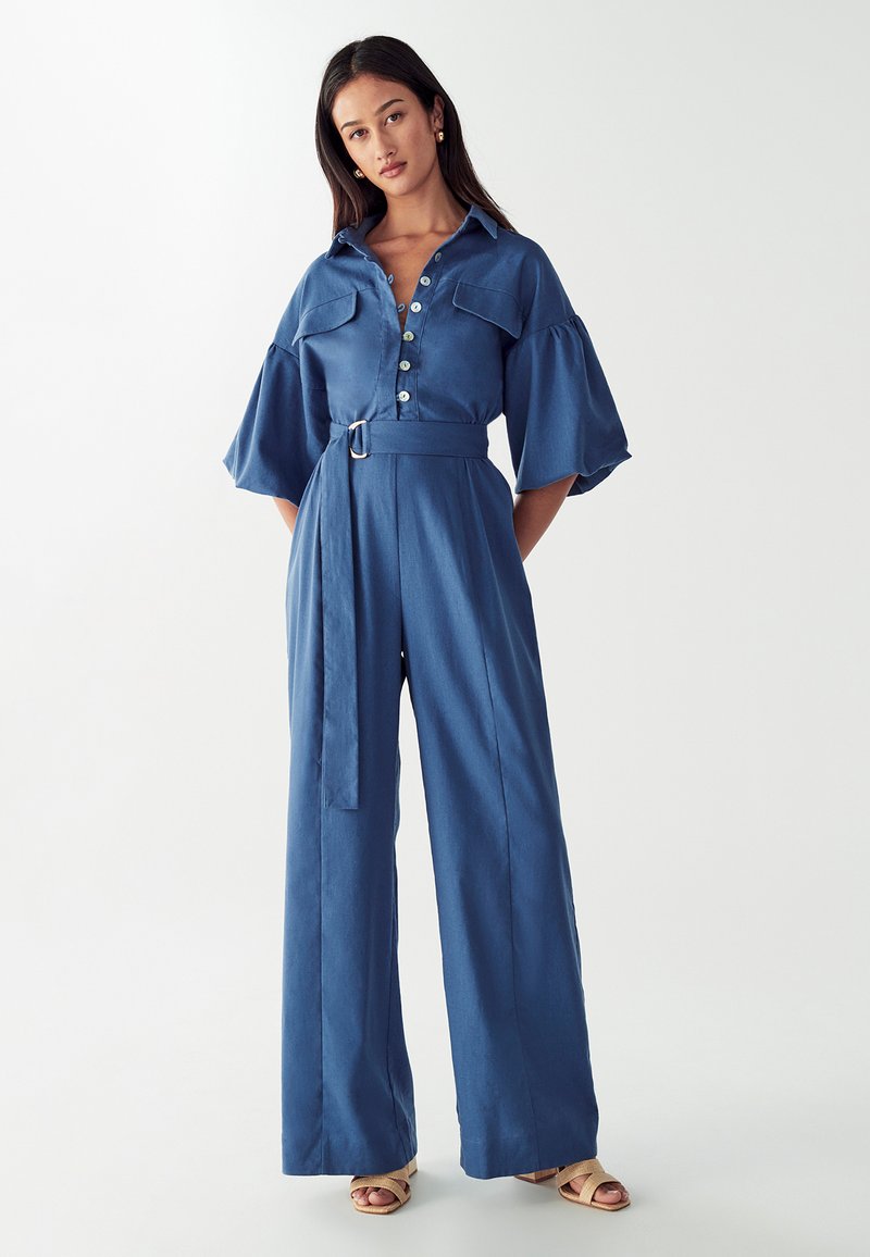 Kvinna i en blå jumpsuit med bälte och vida ben, knapplåst fram och puffärmar, som står mot en enfärgad vit bakgrund.