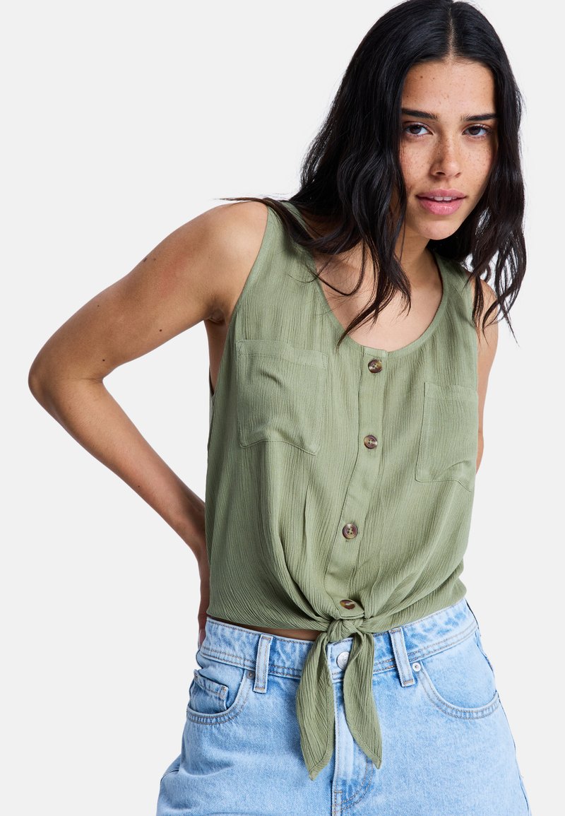 Blusa sin mangas de color verde oliva con nudo en la parte delantera, que presenta dos bolsillos en el pecho y pequeños botones marrones, hecha de tela texturizada.