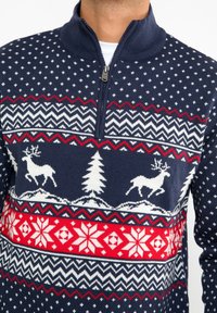 Marineblauer Strickpullover mit einem Reißverschlusskragen, der winterliche Muster aufweist: Rentiere, Bäume und geometrische Designs in weißen und roten Akzenten.