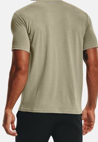 Man som bär en ljus olivgrön Under Armour t-shirt med kort ärm och svarta byxor, visad från ryggen med en tatuering på sin högra arm.