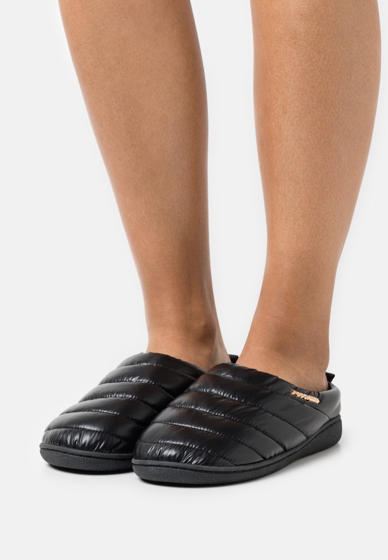 Pieds portant des chaussons d’intérieur noirs matelassés à enfiler avec des semelles épaisses et rembourrées sur fond blanc.