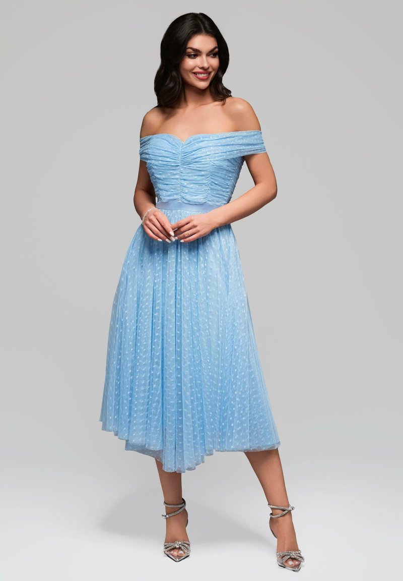 Robe bleu clair à épaules dénudées avec corsage froncé, jupe plissée et pois texturés, associée à des talons argentés à lanières.
