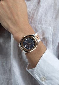 Aštuonkampis laikrodis su tamsiai mėlynu tekstūruotu ciferblatu, sidabriniu ir rožinės aukso nerūdijančio plieno dirželiu. Apsirengtas ant riešo, derinamas su baltu lininio marškiniais.