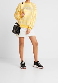 Persona que lleva un sudadera a rayas amarillas y blancas con el texto "Venice Vibes", una falda de mezclilla blanca, zapatillas negras y un bolso negro de bandolera.