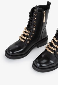 Stivaletti neri in pelle con hardware dorato, caratterizzati da un punta lucida e decorazioni di perline lungo i lacci. Chiusura con zip laterale.