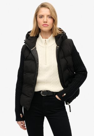 Schwarze Pufferweste mit Kapuze, ausgestattet mit einem strukturierten Strickärmel. Getragen über einem cremefarbenen Kabelstrickpullover, ergänzt durch schwarze Hosen und einen Gürtel.