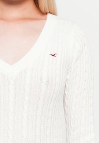 Close-up de uma pessoa a usar um suéter branco com padrão de trançado em V, apresentando um pequeno logotipo de um pássaro vermelho no peito.