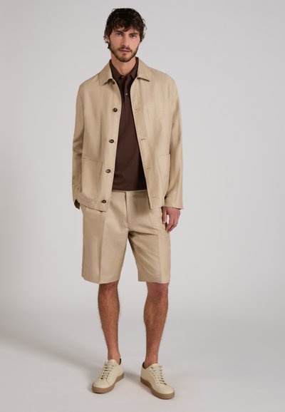 Man standing wearing beige buttoned jacket, matching knee-length shorts, brown polo shirt, and beige sneakers on a plain background.
