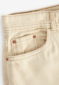 Beige katoenen twill stof met diagonale ribbels, voorzien van een kleine patch-pocket en koperen klinknagels aan de rand van de zak.