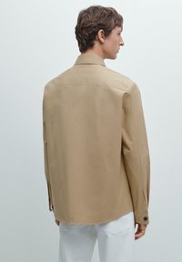 Camicia button-up color marrone chiaro con taglio dritto, maniche lunghe e colletto. Presenta un tessuto liscio e texture, con orlo arrotondato.