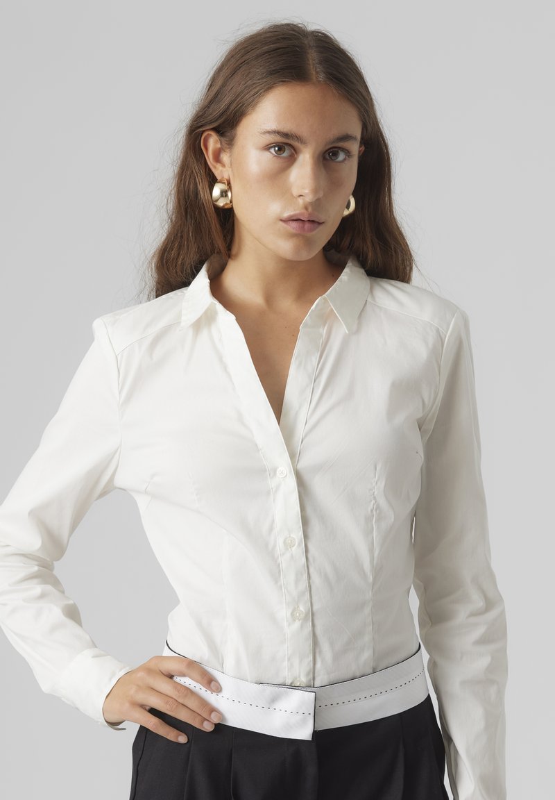Chemise blanche à col, à manches longues, avec une fermeture à boutons à l'avant et une texture lisse. Assortie à un pantalon noir et une ceinture contrastante.