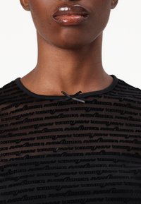 Svart gjennomsiktig topp med teksturert design og gjentatt "Tommy Jeans" logo. Den har rund hals og en dekorativ sløyfe ved kragen.