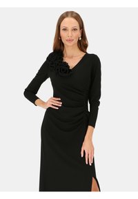 Czarna sukienka z dekoltem w serek, długimi rękawami i marszczeniami po bokach. Zdobiona materiałowym kwiatem na lewym ramieniu oraz rozcięciem po boku.