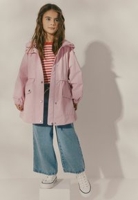 Giacca impermeabile rosa con cappuccio, due tasche frontali e dettagli con bottoni a pressione, abbinata a jeans a gamba larga e sneakers bianche.