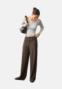 Žena si upravuje hnědé široké kalhoty s vysokým pasem, má na sobě šedý žebrovaný top s odhalenými rameny, černé boty a drží černou tašku s mačkaným designem.