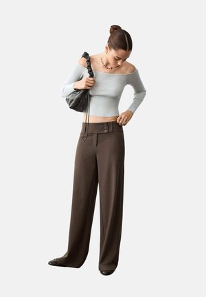 Frau justiert hoch taillierte braune weite Hose, trägt schulterfreies hellgraues geripptes Oberteil, schwarze Schuhe, hält schwarze geraffte Handtasche.