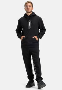 Felpa nera con cappuccio, tasca frontale e grafica bianca. Abbinata a pantaloni della tuta neri con stampa del logo laterale e sneakers coordinate.