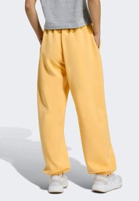 Osoba v voľných žltých teplákoch, šedej košeli a bielych teniskách, stojí s rukami v vreckách na jednolatom pozadí.