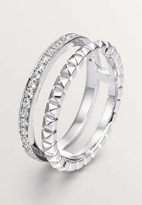 Dos anillos de plata apilados uno sobre otro; uno presenta piedras preciosas claras en un engaste de garra, mientras que el otro tiene un diseño geométrico texturizado.