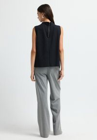 Femme se tenant de dos, portant un haut noir sans manches avec une fermeture éclair au dos et un pantalon gris ample, devant un fond blanc uni.