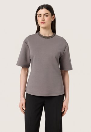 Šedá košile s krátkým rukávem, s texturovaným límečkem ozdobeným korálky, kulatým kolárem, volným střihem a hladkým materiálem. V kombinaci s černými kalhotami.