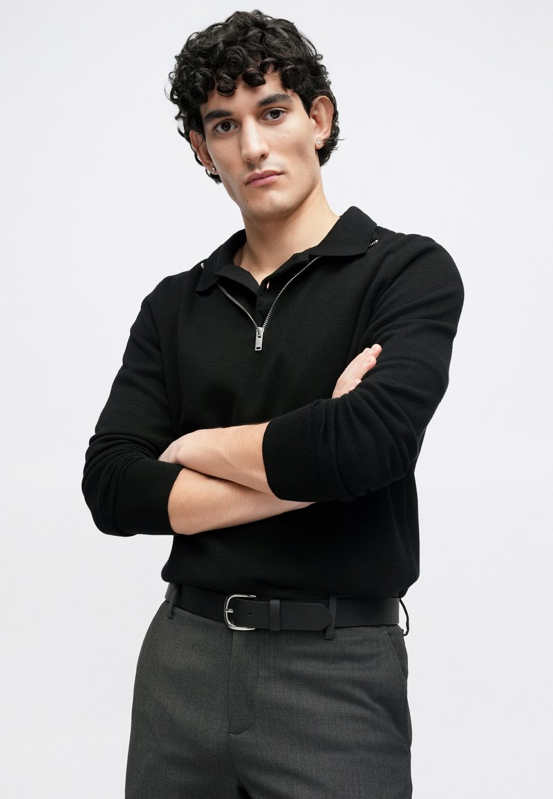 Black zip-up polo sweater with a collar, long sleeves, and a textured finish. Paired with grey trousers and a black belt.