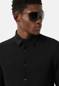 Schwarzes Langarmhemd mit einem strukturierten Kragen, versehen mit einer Knopfleiste und glatter Oberfläche. Das Modell trägt übergroße, dunkle Sonnenbrillen.