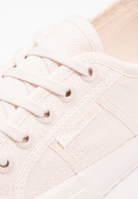 Gros plan d'une basket beige en toile avec des lacets blancs, des œillets métalliques et une étiquette en tissu Superga sur le côté.