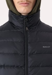 Black puffer vest with a high collar, zipper closure, and visible stitching. Features a logo on the left chest and a green hood underneath.