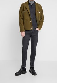 Olivgrön texturerad jacka med stora vita knappar, grå skjorta, mörkgrå slim fit-jeans och svarta snörskor.