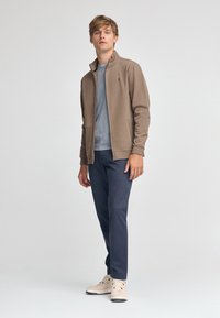 Giacca in fleece con zip di un caldo marrone, abbinata a una t-shirt azzurro chiaro, pantaloni blu scuro e sneakers color crema. Design versatile e funzionale.