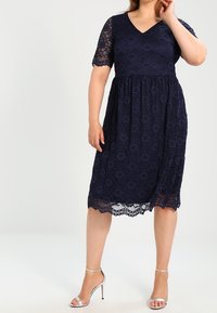 Granatowa sukienka koronkowa z dekoltem w kształcie V, krótkimi rękawami i rozkloszowanym dołem. Ma kwiatowe wzory oraz falbaniasty brzeg. Połączona z srebrnymi szpilkami.